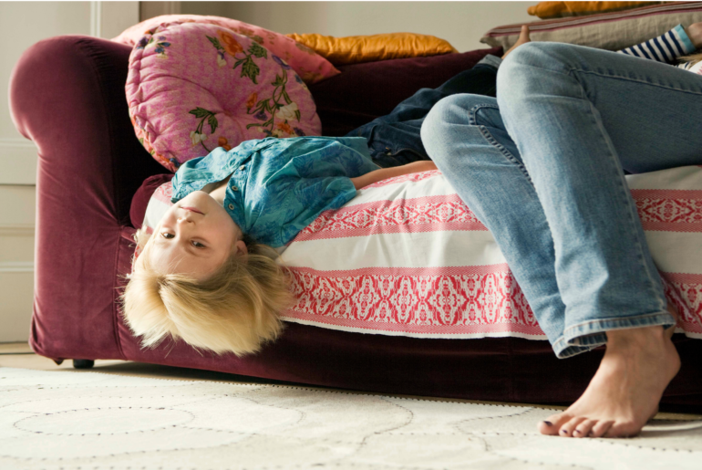 Bored child hanging their head off of the couch as their parent sits next to them.
