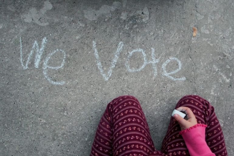 Child writing "We Vote" in chalk.