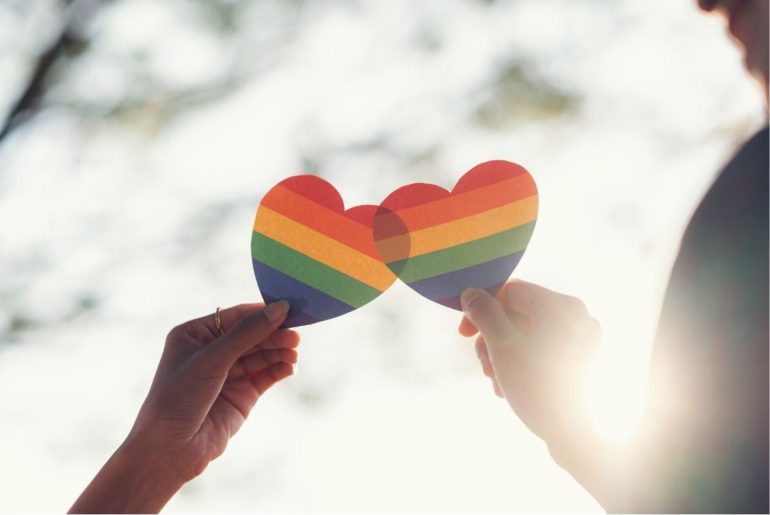How to support an LGBTQ+ kid. Two people holding Pride hearts with sun in background.
