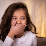 Kid with long dark brown hair covers mouth as if she just said or did something in error