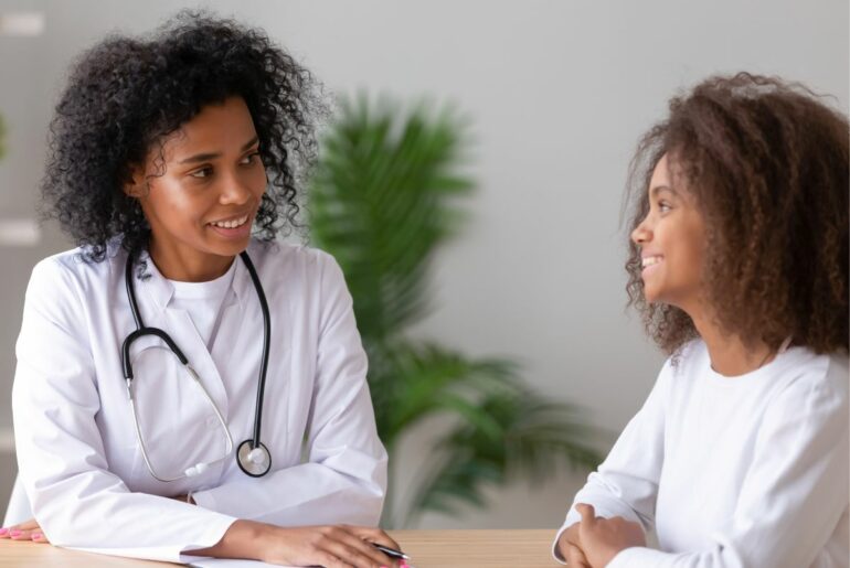 A doctor in white coat, brown skin and curly black hair smiles at a teen with white shirt, brown skin and curly black hair