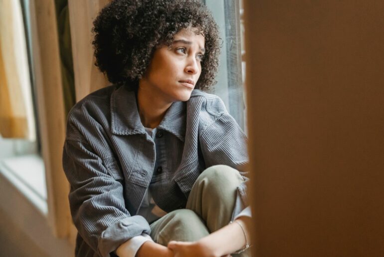 Woman with curly black hair, brown skin and a sad facial expression looking out the window