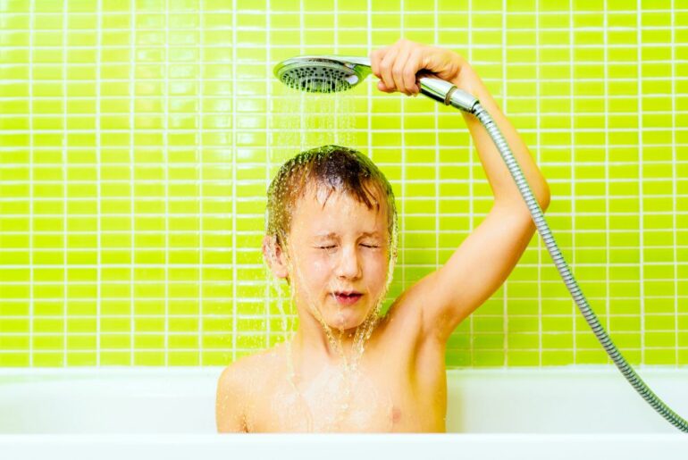 A light-skinned boy holds a shower head over his head, eyes closed, in front of a bright green tile wall