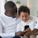 A Black dad and child sit on the couch together, looking at a smartphone and smiling