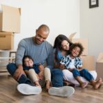 A multiracial family of four sits on a bare wooden floor, with moving boxes all around them